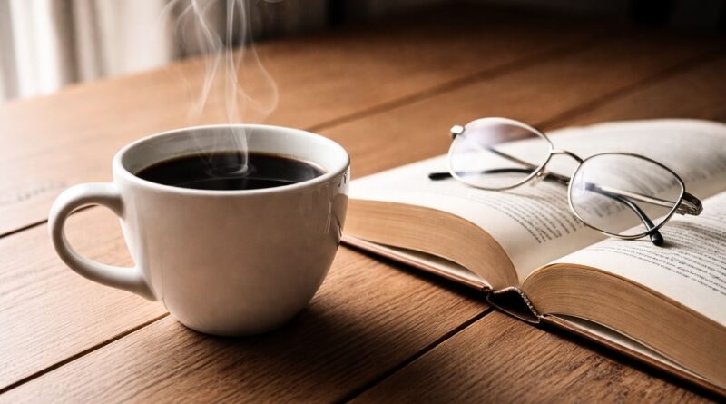 “A steaming cup of coffee on a wooden table beside an open book, representing how coffee enhances focus, comfort, and daily enjoyment”
